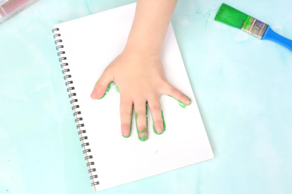 Letter F Frog Handprint Craft - Cultivating Brilliant Minds Child's hand smears bright green paint on a white spiral notebook for this handprint art