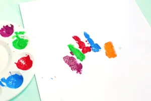 White sheet with multicolored fingerprint paint marks (blue, green, red, purple, orange) near a paint palette on the left side for this back to school craft
