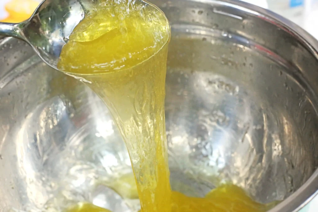 Lemon Slime - Cultivating Brilliant Minds Close-up of a thick yellow liquid being poured from a spatula into a stainless steel bowl, creating a viscous stream.