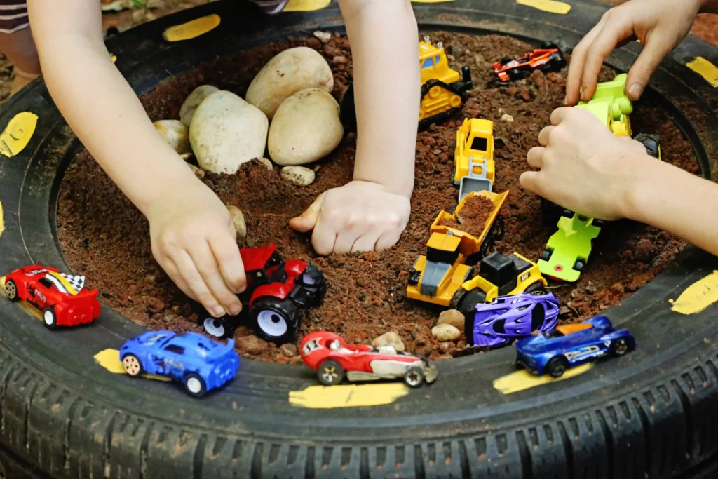 Children's hands arranging colorful toy cars inside a dirt-filled circular tire play pit with rocks nearby.
