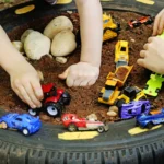 Children's hands arranging colorful toy cars inside a dirt-filled circular tire play pit with rocks nearby.