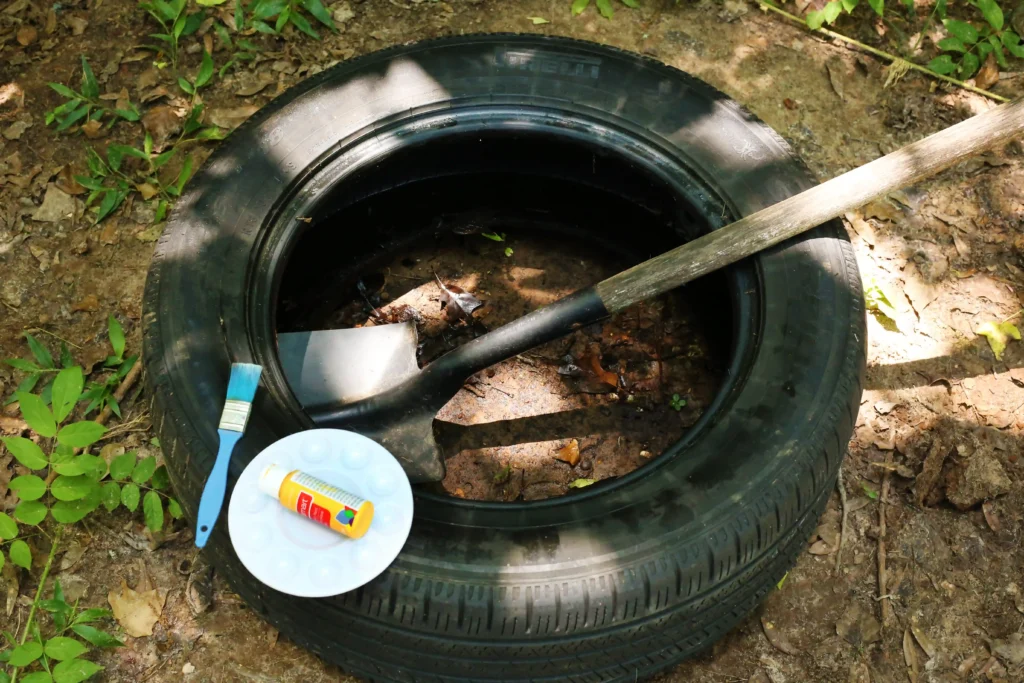 supplies for this outdoor play idea- Discarded black tire on dirt with leaves and scattered painting supplies (a blue-handled brush and a tube) nearby.