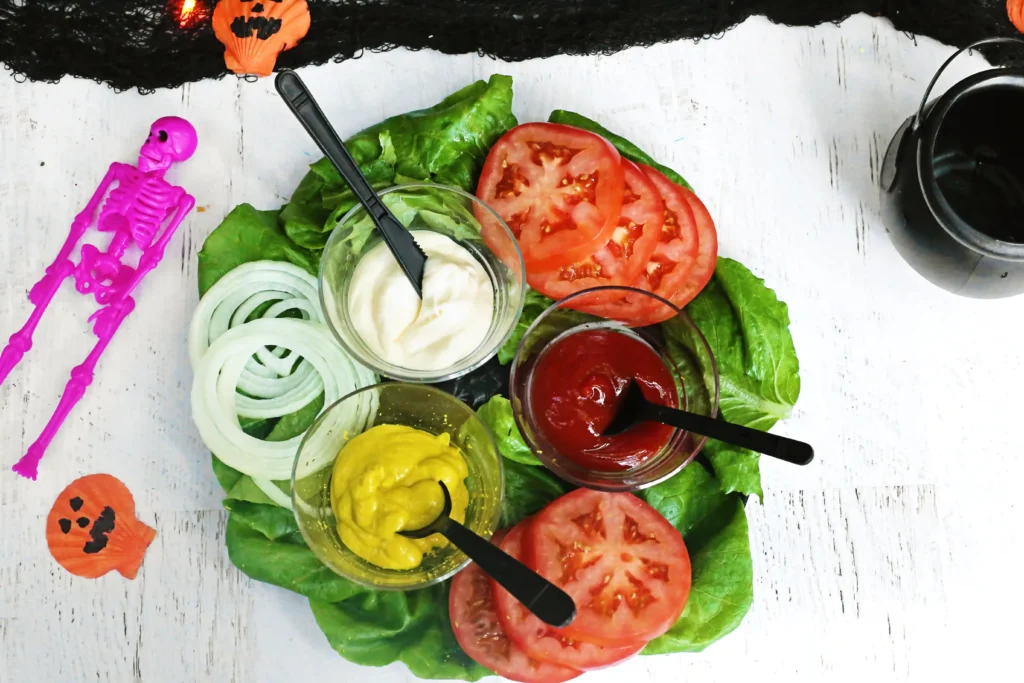 Overhead shot of a colorful salad with tomato slices, lettuce, and onion rings in bowls, beside a pink skeleton toy and Halloween decor on a white table.