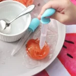 Close-up of a child’s hand squeezing a blue silicone dropper over a plate with frozen green and pink ice and a white cup nearby with a spoon inside it.