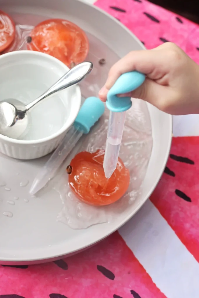 Close-up of a child’s hand squeezing a blue silicone dropper over a plate with frozen green and pink ice and a white cup nearby with a spoon inside it.