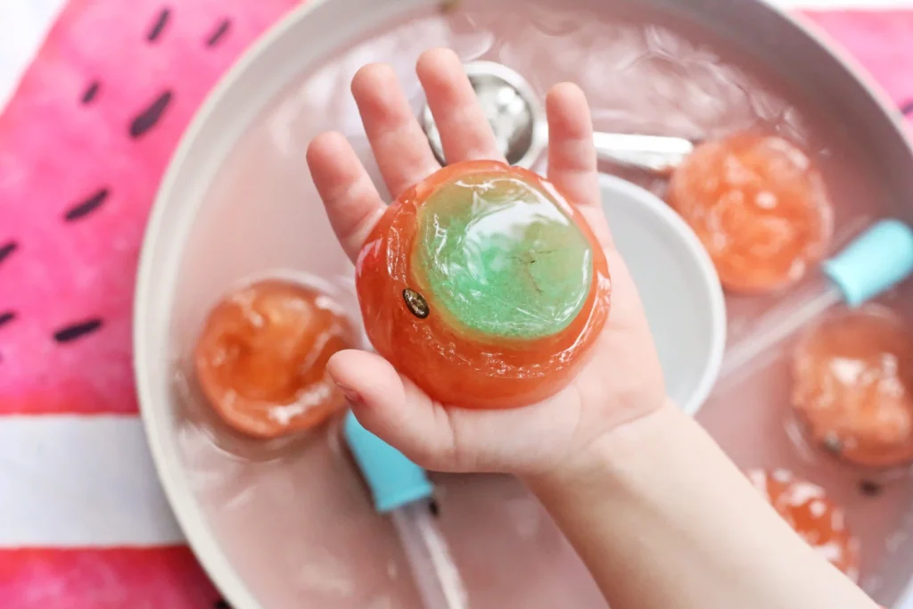 a Watermelon Ice Sensory Play that has a kid holding frozen water dyed pink and green with watermelon seeds