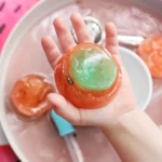 a Watermelon Ice Sensory Play that has a kid holding frozen water dyed pink and green with watermelon seeds