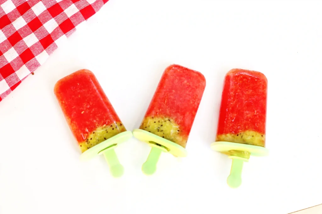 Watermelon Kiwi Popsicles - Cultivating Brilliant Minds Watermelon popsicles on white background with a red and white checkered napkin nearby, pastel green sticks visible.