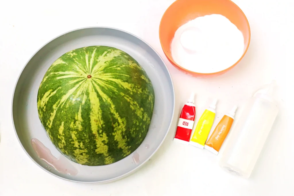 supplies for this summer STEM activity - Watermelon in a gray bowl next to a cup of milk and colorful paint tubes on a white surface.