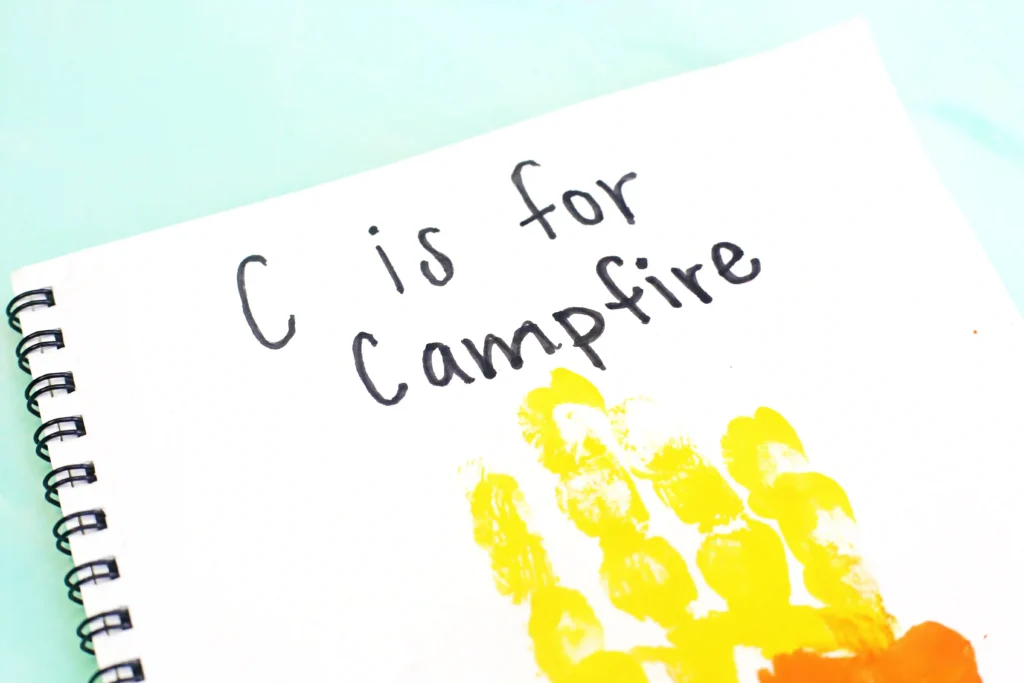 Close-up of a spiral notebook page with handwritten phrase'C is for Campfire' and yellow, orange, and red hand paint stamp designs on white paper.