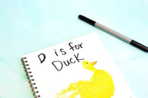Notebook page with handwritten 'D is for Duck' and a yellow duck illustration on a light blue background, with a pen nearby for this alphabet craft for kids