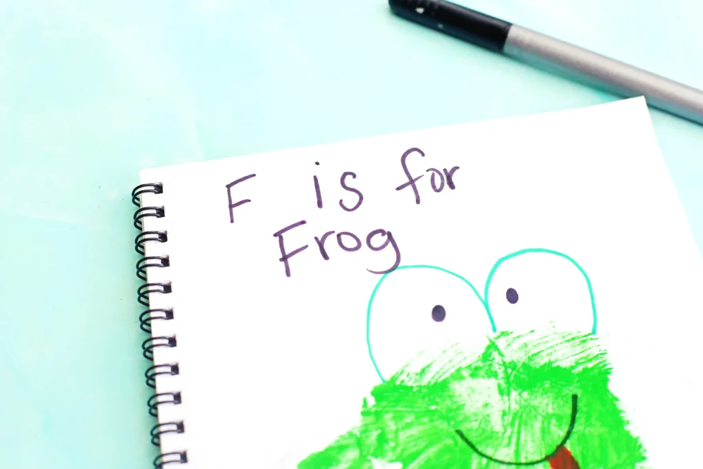 Letter F Frog Handprint Craft - Cultivating Brilliant Minds Notebook page with a green frog illustration and the text'F is for Frog' written in black ink on a mint background, showing a spiral binding on the left for these handprint animal crafts