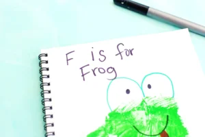 Notebook page with a green frog illustration and the text 'F is for Frog' written in black ink on a mint background, showing a spiral binding on the left for these handprint animal crafts