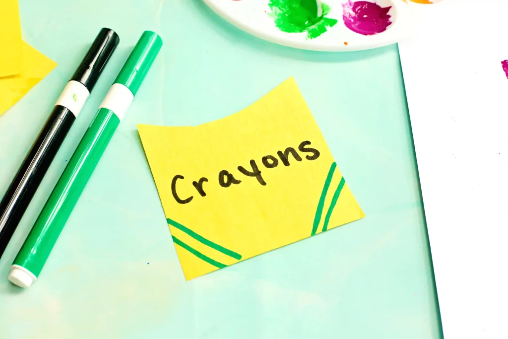 Yellow construction paper cut into the shape of a crayon box with "crayons" written on it on a light blue workspace with scissors and a craft knife nearby.