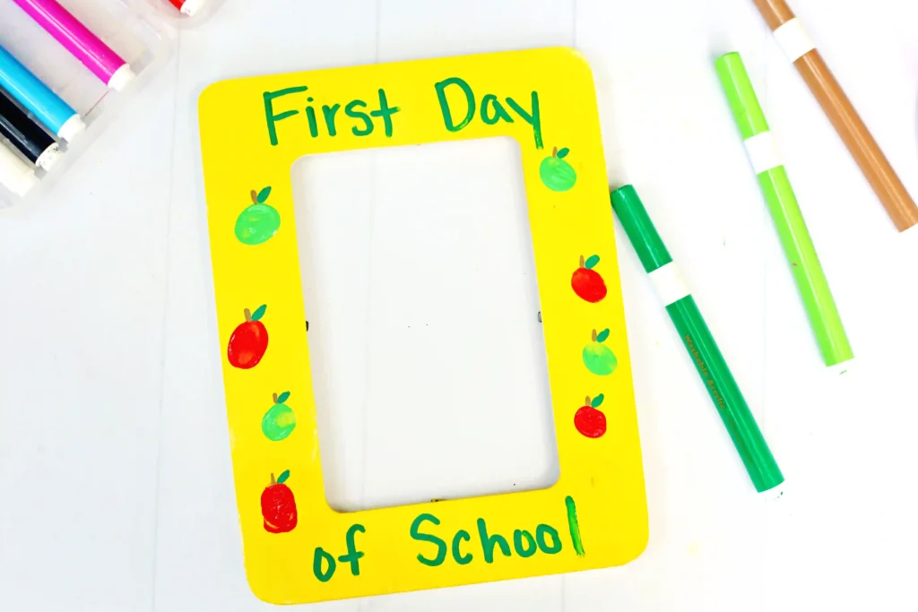 a First Day of School Photo Frame that is painted yellow with fingerprint apples and words that read "first day of school"