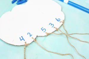 a piece of cardboard cut into the shape of a cloud and painted white with twine tied on and numbers written on the cardboard above the twine