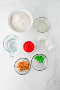 ingredients for these fall cupcake recipes- Top-down view of baking ingredients: bowl of flour, glass of water, eggs, orange pretzel sticks, and green candies on a marble surface.