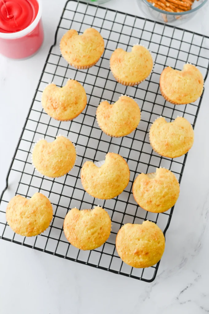 Apple Cupcakes - Cultivating Brilliant Minds Golden muffins cooling on a black wire rack with a hint of pink frosting container at the edge.