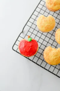 Red-frosted cupcake with a small green candy on top on a black wire cooling rack, with vanilla cupcakes nearby in the background for these back to school desserts