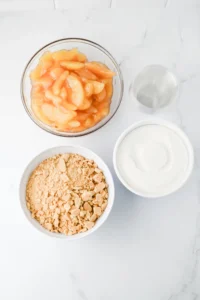 ingredients for this classroom treat - Top-down view of apple slices in a glass bowl, crushed graham crackers in a white bowl, and cool whip in another bowl with a glass of water on a white marble surface.