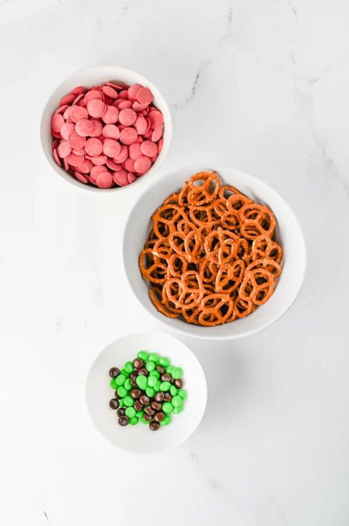 ingredients for this back to school treat -Overhead view of white bowls filled with pink candy coins, orange pretzels, and green and brown chocolate candies on a white marble surface.