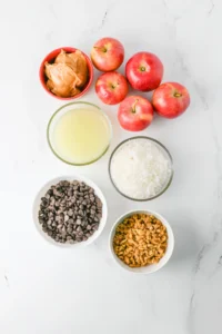 ingredients for the apple cookies- Top-down view of several red apples, peanut butter in a red bowl, a glass of pale liquid, rice, chocolate chips, and granola on a white marble surface, ready for a snack bowl recipe.
