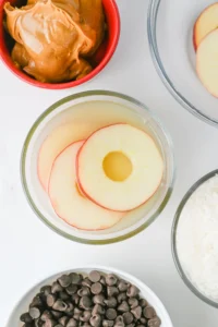 Overhead view of bowls with sliced apples on yogurt and a dollop of peanut butter nearby, plus chocolate chips in a separate bowl for this no-bake snack.