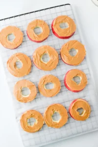Row of donut-shaped apple slices with orange icing cooling on a parchment-lined grid for these classroom snacks