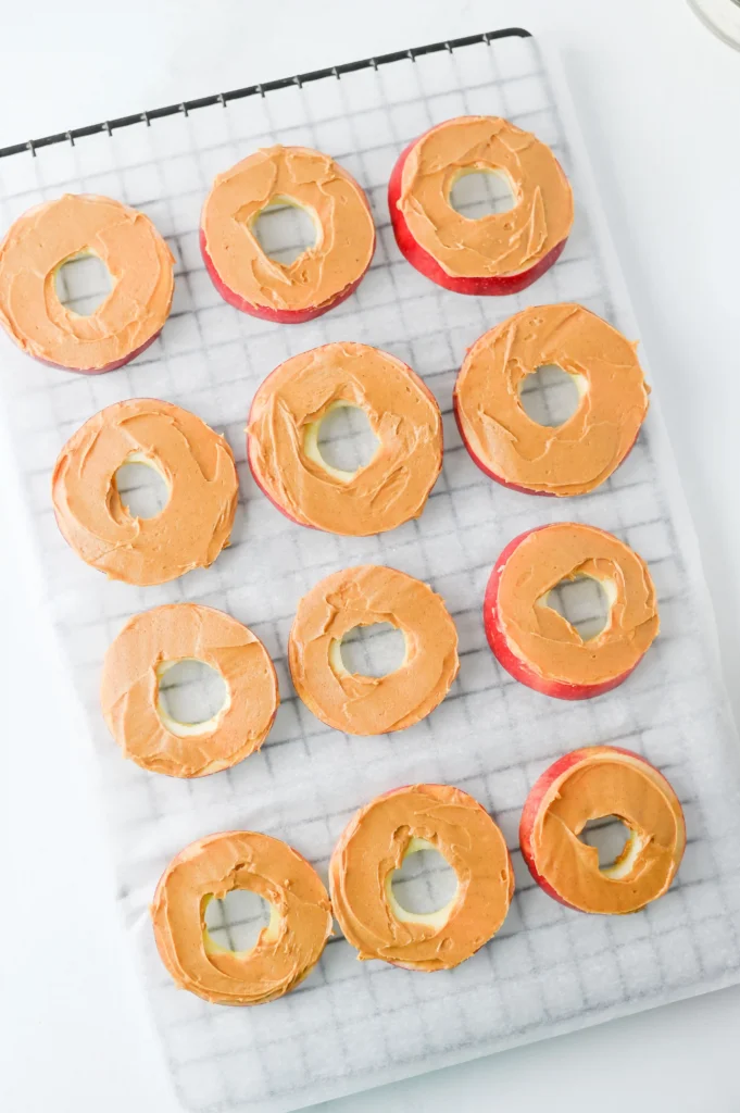 Row of donut-shaped apple slices with orange icing cooling on a parchment-lined grid for these classroom snacks