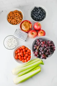 ingredients for these celery bug snacks: bowls of blueberries and orange pretzels, apples, grapes, cherry tomatoes, celery stalks, and a peanut butter container with a cereal snack nearby.