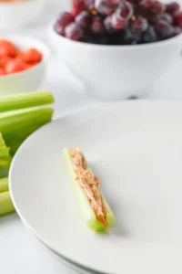 Celery stick topped with peanut butter on a white plate, with a bowl of red grapes blurred in the background.