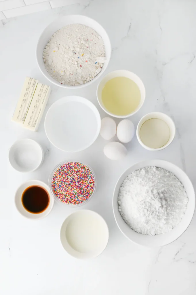 ingredients for this dot birthday cake: bowls of flour, a yellow liquid, eggs, butter sticks, and colorful sprinkles on a white marble surface, ready for dessert mixing
