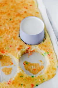 Close-up of a round paper ice cream cup pressed into rainbow sprinkles cake batter in a baking tray.