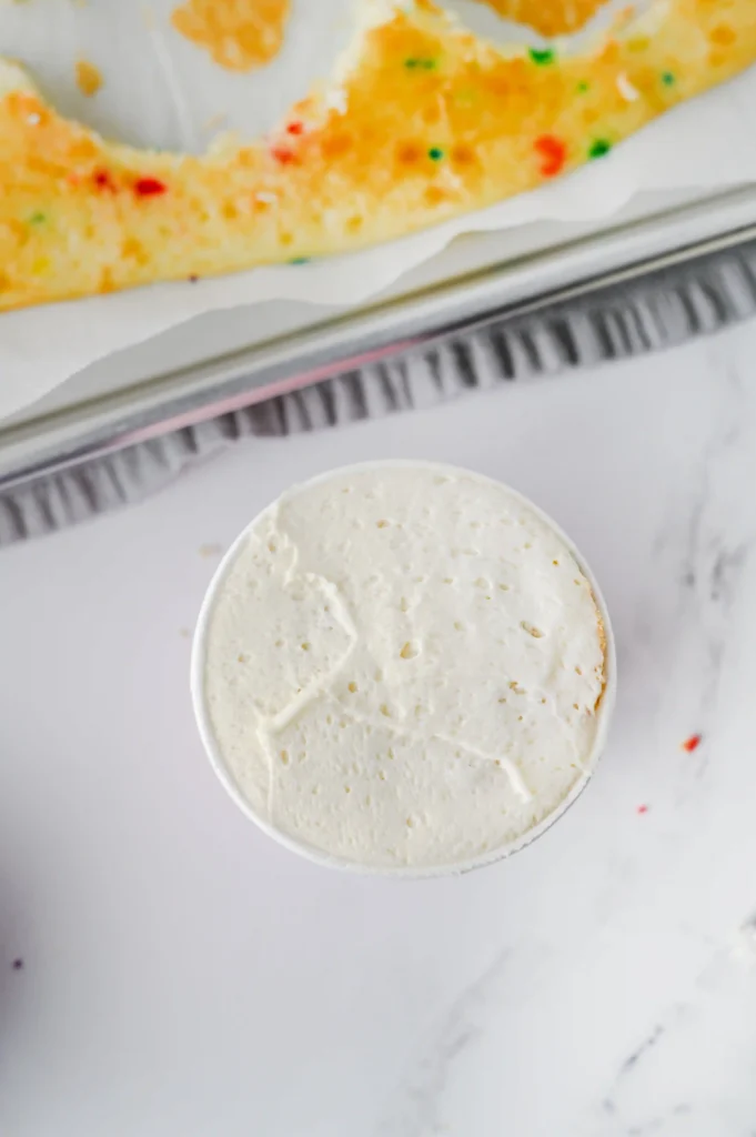 Frosted cake in a paper cup on a marble surface, with a colorful sprinkled pastry blurred in the background.
