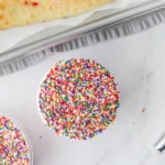 Cupcake topped with multicolored rainbow sprinkles on a white surface, viewed from above.