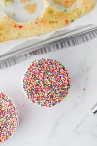 Cupcake topped with multicolored rainbow sprinkles on a white surface, viewed from above.