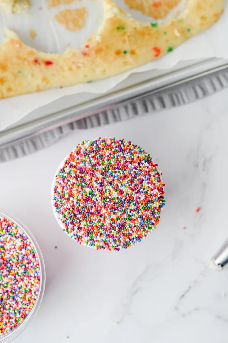 Cupcake topped with multicolored rainbow sprinkles on a white surface, viewed from above.