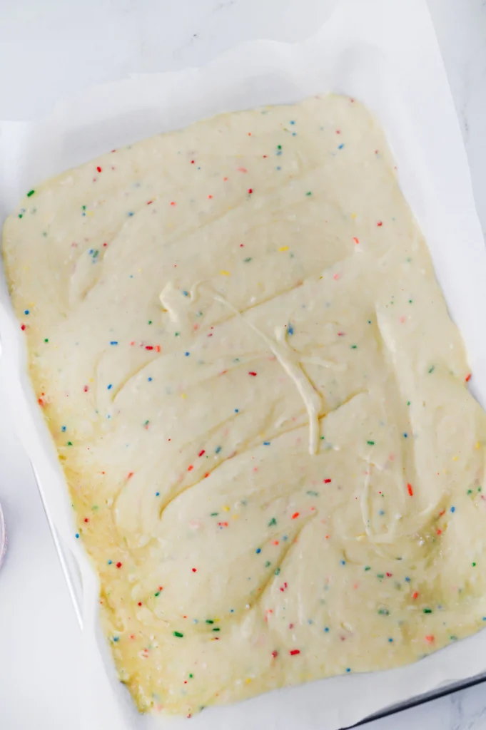 Unbaked funfetti cake dough with colorful rainbow sprinkles spread on parchment paper in a baking dish.