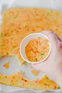 Hand holding a white cup with a bite-sized piece of confetti cake over a large sheet of rainbow-speckled cake on parchment