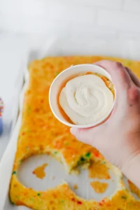 Close-up of a hand applying white frosting from a cup onto a golden, ring-shaped pastry with colorful sprinkles.