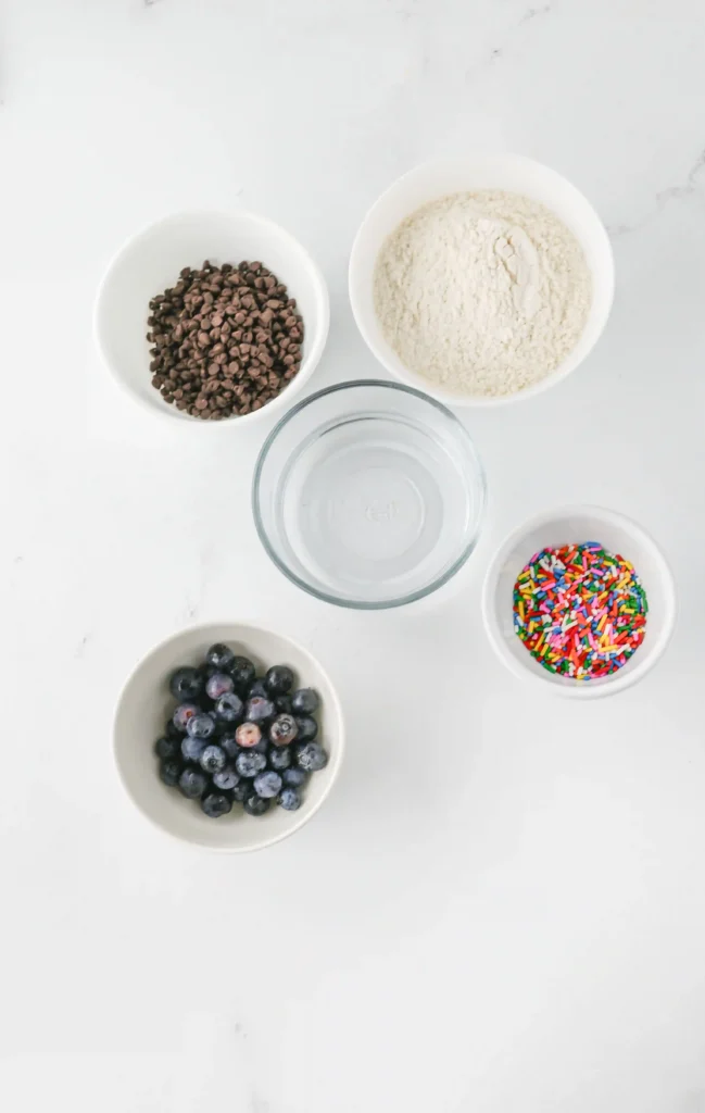 supplies for the back to school breakfast ideas - Overhead view of bowls with chocolate chips, flour, sprinkles, blueberries, and a glass of water on a white surface.