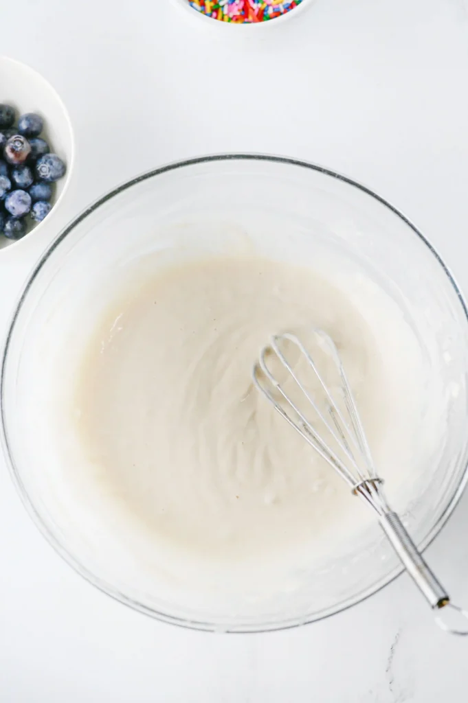 Top-down view of a large glass mixing bowl with a metal whisk inside; a small bowl of blueberries sits to the left and a bowl of colorful sprinkles is above.