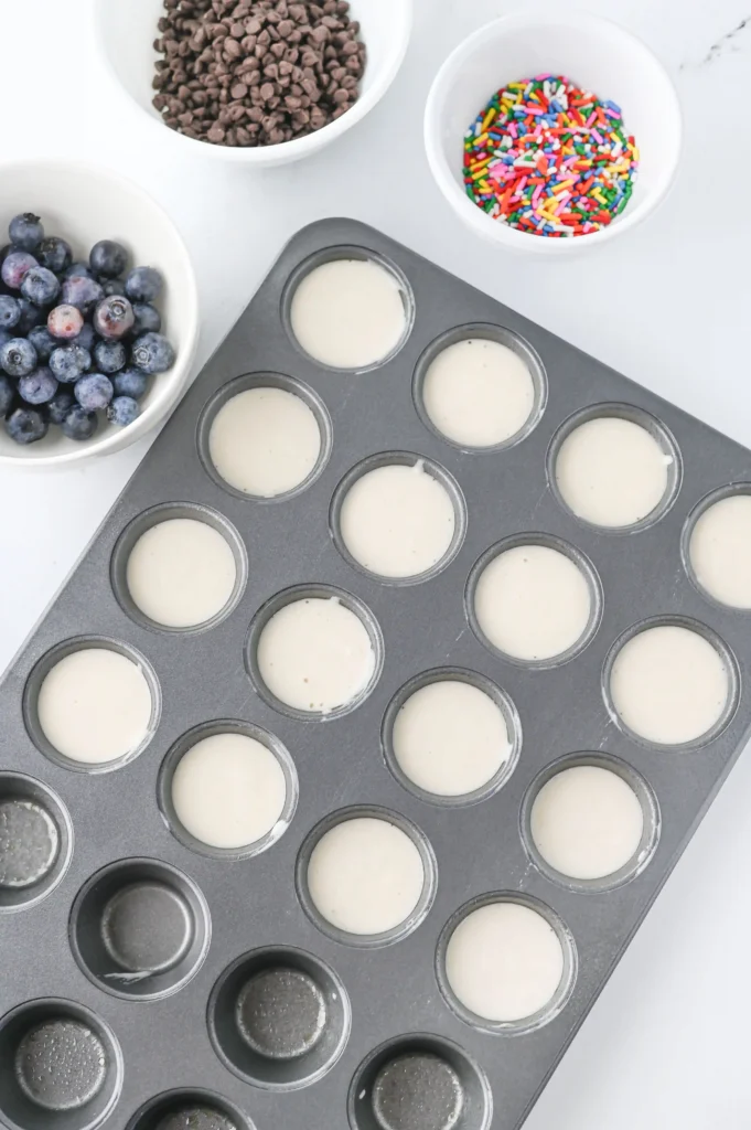 Muffin tin with batter cups, surrounded by bowls of blueberries, chocolate chips, and rainbow sprinkles for toddler breakfast ideas