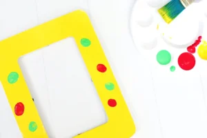 Close-up of a bright yellow frame with green and red paint spots on a white surface, alongside a paint palette and brush for this first day of school activity