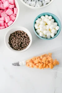 Overhead view of colorful bowls of candy: pink wrapped starburst, white marshmallows, and chocolate chips on a light surface for this back to school snack