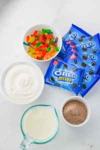 ingredients for this easy snack for kids: Assorted baking scene with Oreo Mini packets, a bowl of colorful gummy worms, bowls of white frosting and cocoa powder, and a pitcher of milk on a marble countertop