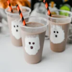 Chocolate Milk Ghost Cups that have marshmallow fluff ghosts in a cup with chocolate milk and an orange and black straw