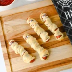 Halloween-themed mummy hot dogs on a wooden board with red ketchup and yellow mustard nearby.