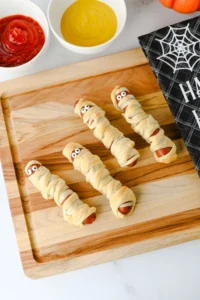 Halloween-themed mummy hot dogs on a wooden board with red ketchup and yellow mustard nearby.