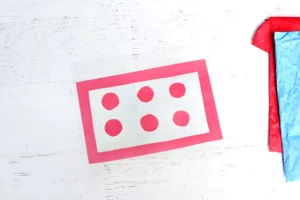 Red rectangular outline with red dots on a white wooden surface, beside red and blue tissue-wrapped gift on the right.